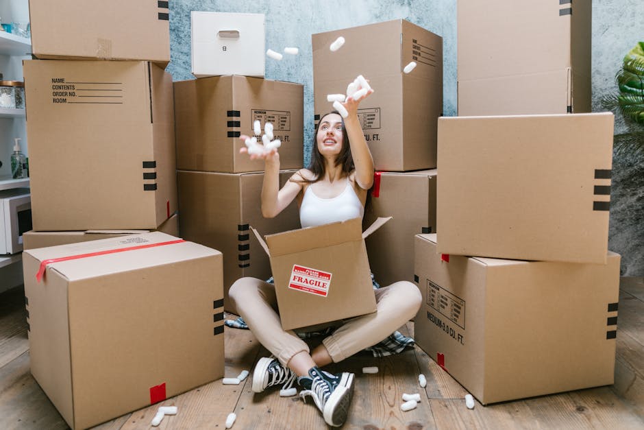 A young woman with long dark hair and casual clothing is sitting on a wooden floor, surrounded by large brown cardboard moving boxes of various sizes, some sealed with tape and marked with labels. She
