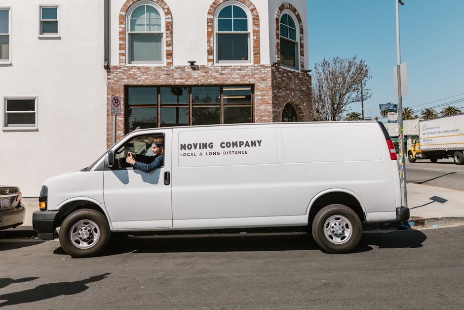A white moving van with the text 'MOVING COMPANY LOCAL & LONG DISTANCE' parked on a city street in front of a brick building with arched windows. The driver, wearing sunglasses and a dark jacket, is s