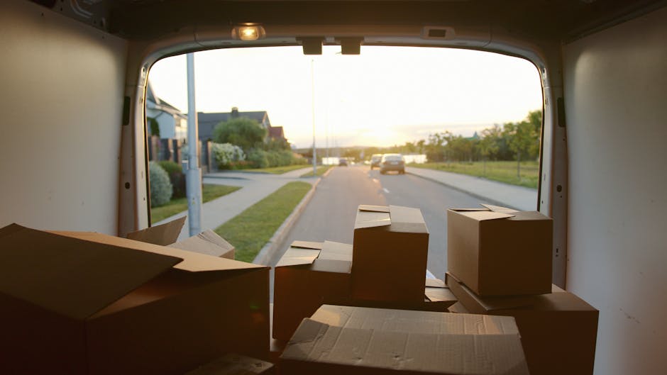 A white moving van with the text 'MOVING COMPANY LOCAL & LONG DISTANCE' parked on a city street in front of a brick building with arched windows. The driver, wearing sunglasses and a dark jacket, is s