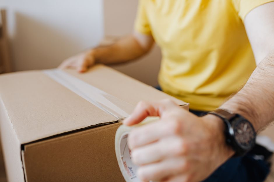 Close-up of a person wearing a yellow t-shirt and a black wristwatch, sealing a cardboard moving box with clear packing tape during a home relocation. The individual is inside a well-lit room, with th