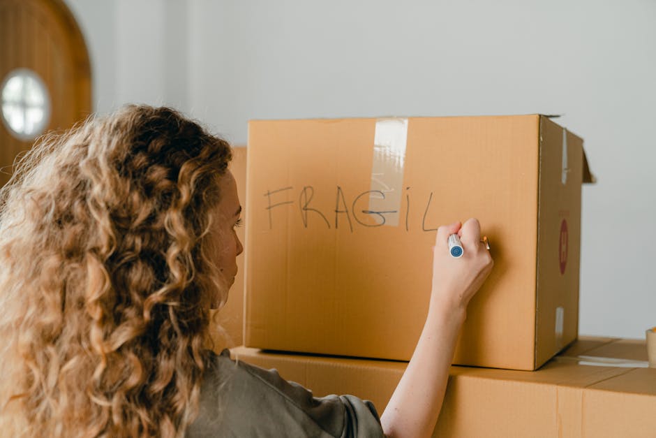 Close-up of a person wearing a yellow t-shirt and a black wristwatch, sealing a cardboard moving box with clear packing tape during a home relocation. The individual is inside a well-lit room, with th
